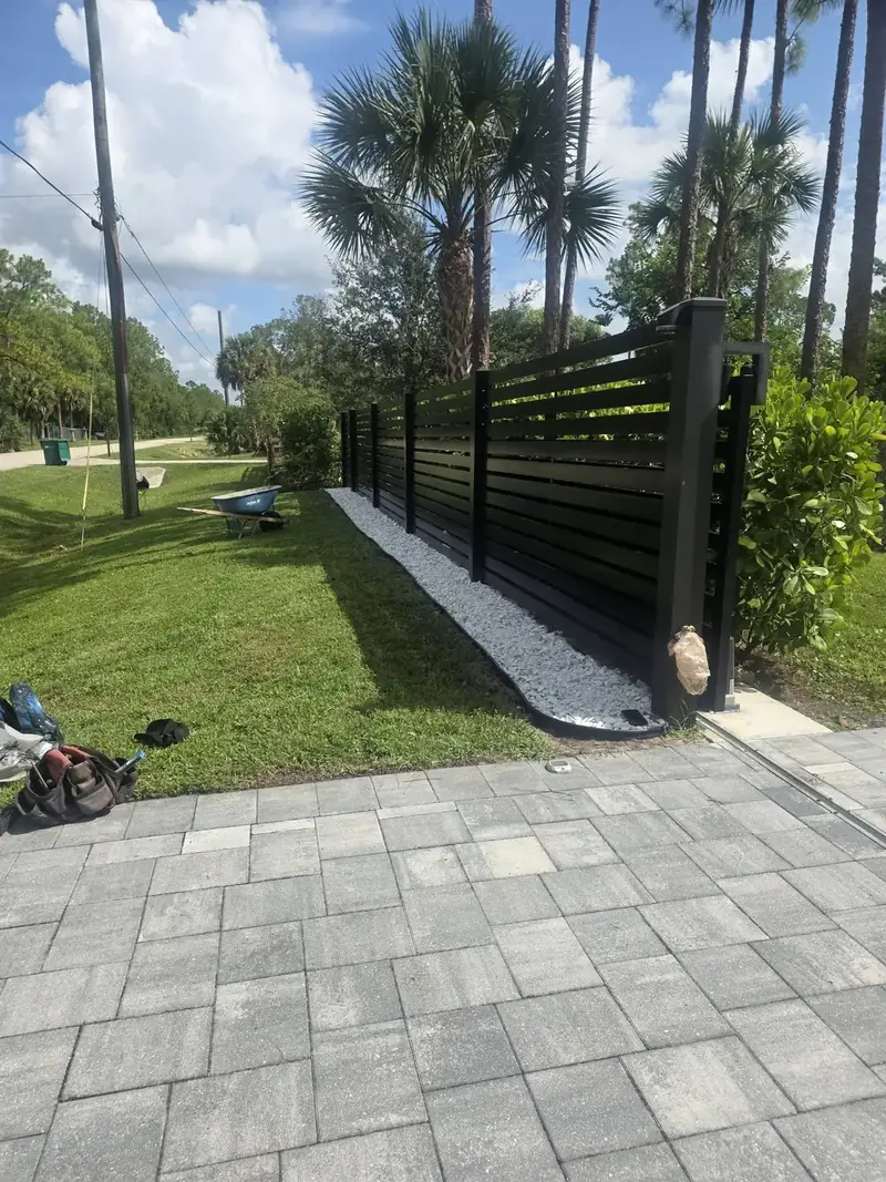 Black horizontal slats run between square posts along the property line, finished with a white-rock border beside the new pavers.
