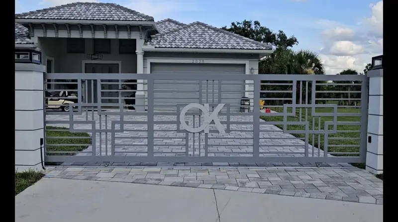 A gray driveway gate with an open geometric grid and a custom monogram spans the paver driveway between white columns topped with lantern lights.