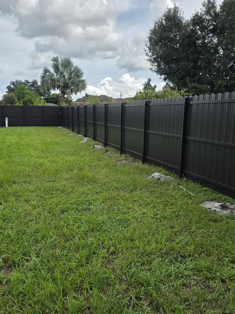 A long dark privacy fence with shaped picket tops follows the edge of a broad lawn beneath a cloudy Florida sky.