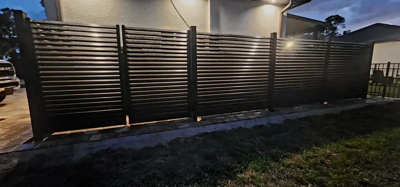 A wide evening view shows dark horizontal privacy panels along the lawn, with lighting beside the home and a white-rock border below.