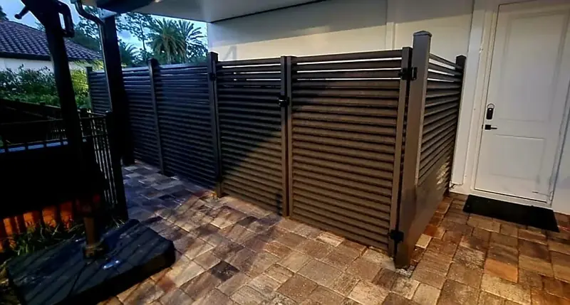 A dark pedestrian gate with closely spaced horizontal slats opens onto a covered paver walkway beside the home.