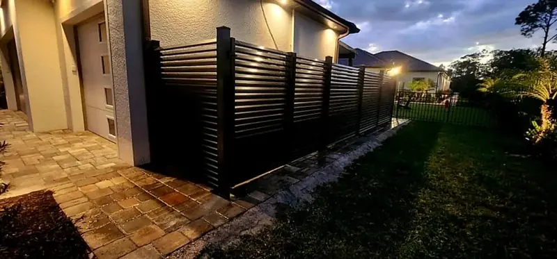 Warm exterior lighting highlights dark horizontal privacy panels and the paver path running along the side of the home.