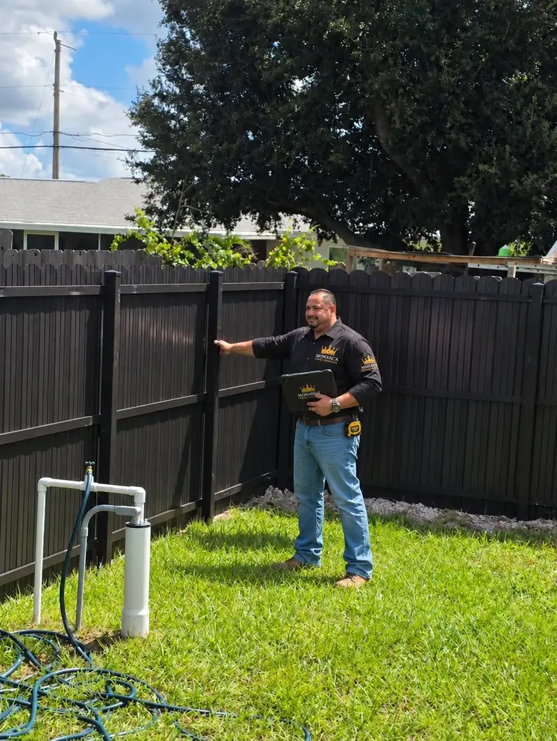 A Monarca team member checks a finished dark privacy fence with picket-style tops and a white-rock strip along its base.