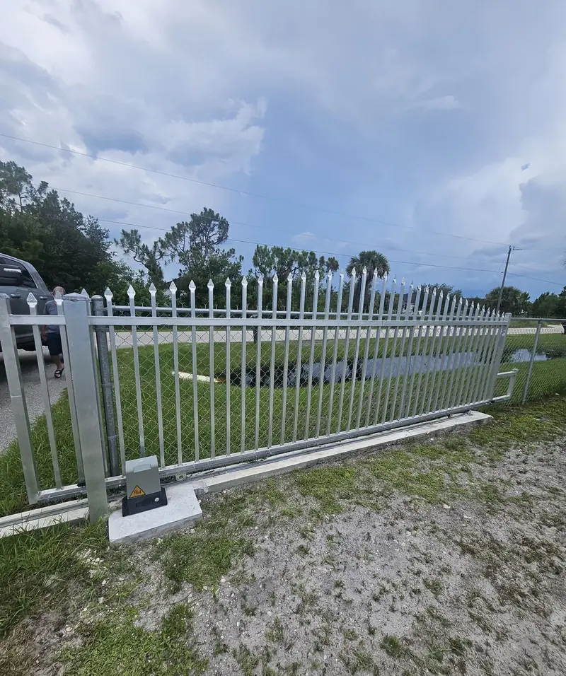 A white picket driveway gate beside an open field and gravel approach.