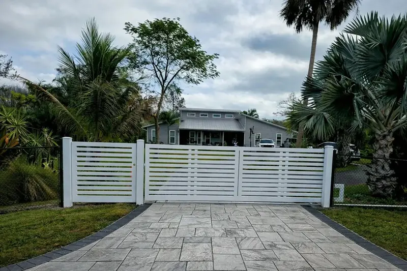 A white double driveway gate with horizontal slats and a matching walk gate were set between square posts at the end of a stone-pattern driveway.